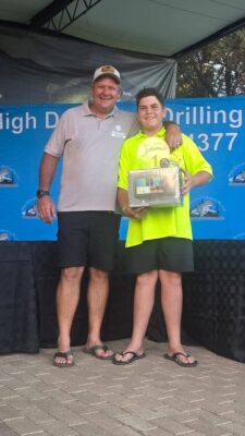 A smiling man in a cap and shorts stands with his arm around a boy in a bright yellow shirt holding a prize. They are in front of a blue banner at an event.