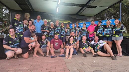 A group of 23 people pose together under a canopy, wearing matching fishing-themed shirts. They are smiling and seated closely, creating a sense of camaraderie.