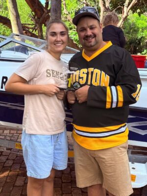 A smiling man and woman stand close, holding an award together. The man wears a Boston sports jersey, the woman a casual shirt, with a boat and trees in the background.