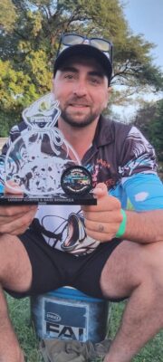 A man in a colorful fishing shirt sits on a bucket, smiling and holding a trophy that reads "Loskop Kurper & Bass Bonanza 2023". Trees are in the background.