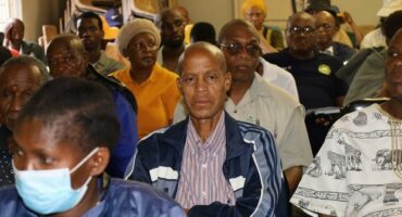 A diverse group of people attentively listen in a meeting room. The focus is on a man in a striped shirt and blue jacket. The atmosphere is thoughtful and serious.