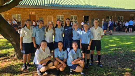 A group of ten students in uniform pose with a teacher under a tree outside a brick school building. The scene is sunny, conveying a cheerful mood.