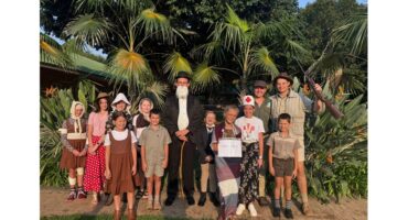 A group of children and adults are in costume outside in front of tropical plants. They wear vintage and thematic outfits, smiling, conveying joy and creativity.