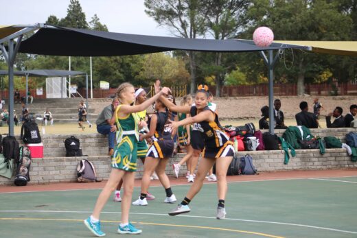 Girls playing netball on an outdoor court. One player in green passes the ball while a player in black defends. Spectators and bags are visible.
