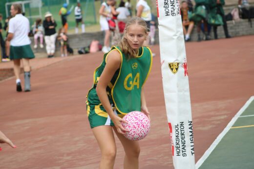 Young girl prepares to throw a pink netball on an outdoor court. She wears a green bib marked "GA" and appears focused. Other players are seen in the background.