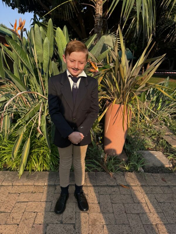 A young boy in a suit stands smiling on a sunlit path, surrounded by lush tropical plants and a large terracotta pot, creating a formal, cheerful atmosphere.