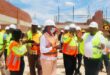 A group of construction workers and supervisors in hard hats and reflective vests discuss plans on a sunny construction site, surrounded by brick walls.