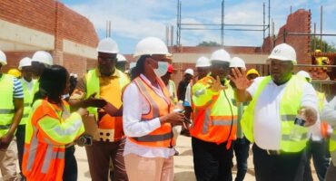 A group of construction workers and supervisors in hard hats and reflective vests discuss plans on a sunny construction site, surrounded by brick walls.