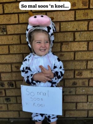 A smiling child wearing a cow costume with a pink cow head hood stands against a brick wall. A sign hangs around their neck reading "So mal soos 'n koei."