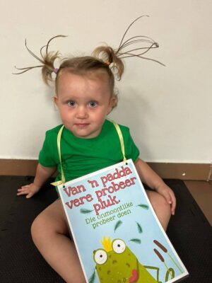 A young child with playful, spiky pigtails sits on a mat wearing a green shirt. They hold a sign with a frog illustration and Afrikaans text. The mood is cheerful and whimsical.