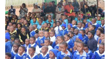 A large group of young schoolchildren in bright blue uniforms sit together, attentively watching an event. Adults are seated behind them, some in colorful attire, creating a vibrant and lively atmosphere.
