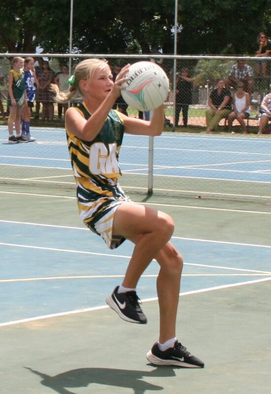 A young athlete in a green and yellow uniform leaps to catch a netball on an outdoor court. Spectators sit watching in the background. Energetic and focused.