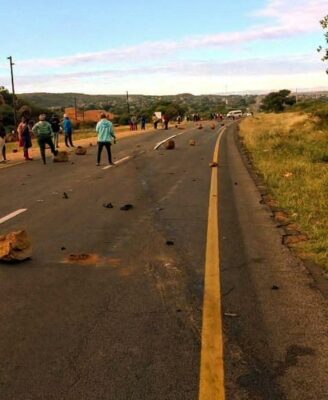 A rural road is blocked by scattered rocks, with people standing on both sides. The scene suggests a protest or obstacle, under a clear sky.
