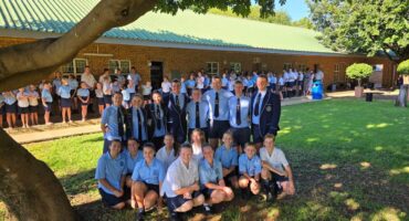 A large group of students in uniforms gather on a sunny lawn. Some stand, while others kneel under a tree, smiling, suggesting a celebratory mood.