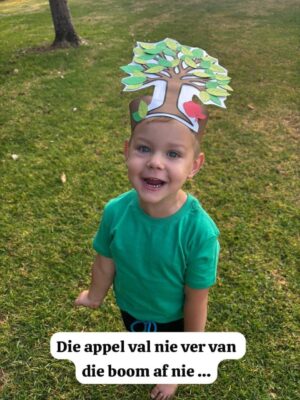 A smiling child on grass wearing a tree-shaped paper hat with leaves and an apple. Text in Afrikaans reads, "Die appel val nie ver van die boom af nie."