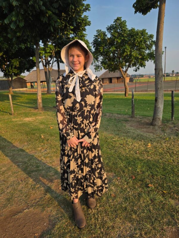 Young person smiling in a floral dress and bonnet, standing on grass with trees and rustic buildings in the background, under a clear blue sky.