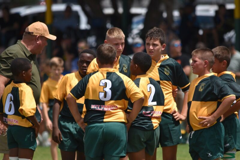 A youth rugby team in yellow and green jerseys, numbered 2, 3, and 6, huddles around a coach during a break. The tone is focused and determined.