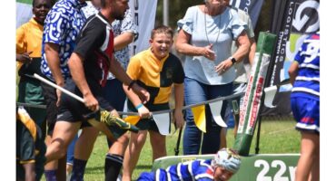 A young rugby player in blue and white stripes falls on the grass as spectators cheer enthusiastically. One boy in yellow and black stands excitedly near the field. The scene conveys energy and support, with people wearing casual summer attire.