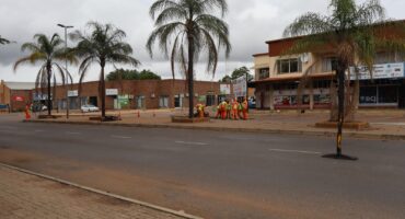 Workers in orange vests gather near palm trees lining a quiet street. The background features shops and cloudy skies, creating a calm urban scene.