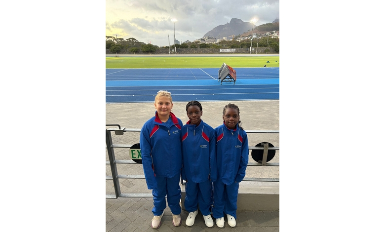 Three children in matching blue tracksuits stand at a sports stadium. A running track and mountain are in the background under a cloudy sky.