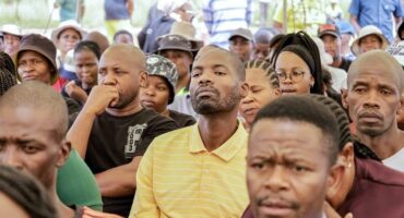A diverse crowd sits attentively at an outdoor gathering, many appear focused and thoughtful. The setting suggests a community meeting or event.
