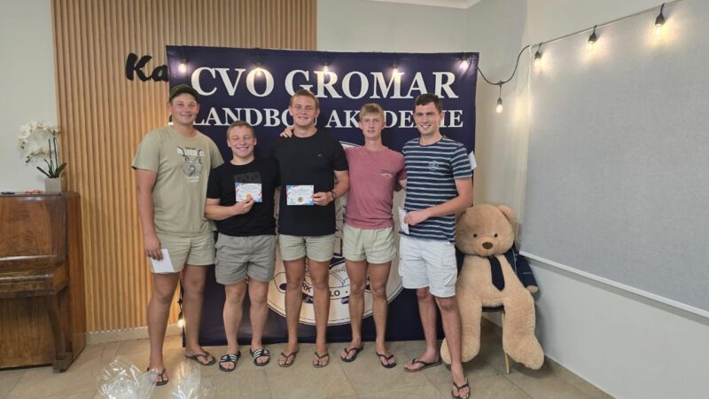 Five young men stand smiling in front of a sign that reads "CVO Gromar." They hold certificates, exuding pride and camaraderie in a casual setting.