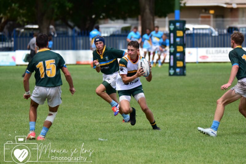 A rugby player in green and yellow stripes holds the ball, evading opponents in a match on a grassy field. Background shows teammates and spectators.