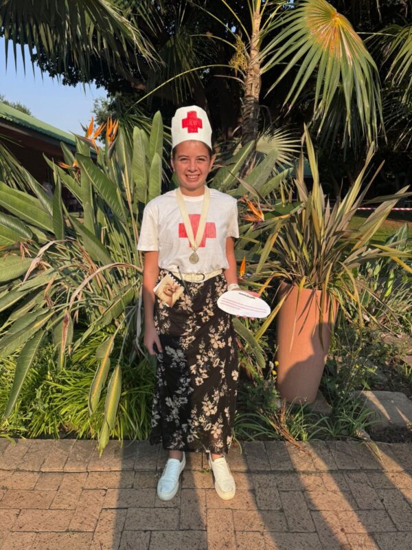 Young person smiling in a nurse costume, wearing a white shirt with a red emblem and paper hat, standing on a patio with lush tropical plants.