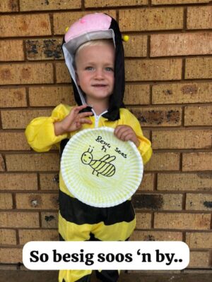 A child in a bee costume smiles, holding a paper plate with a hand-drawn bee that reads "Besig soos 'n by." Stands before a brick wall. Happy tone.