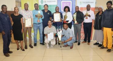 A group of people stand indoors, smiling, holding trophies and certificates. Three banners are displayed behind them, suggesting a celebratory event.