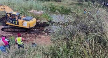 A yellow excavator works near a small pond in a grassy, rural area. Three people in bright clothing observe from the foreground. The scene is calm and natural.