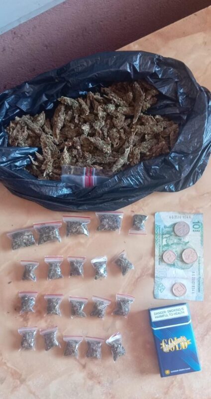 A large black bag filled with dried cannabis is on a table. Below it, there are several small plastic bags, some currency, and a pack of cigarettes.