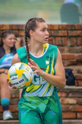 A woman in a green sports uniform holds a netball, focused on a game. A brick wall and a teammate sitting in the background are visible.