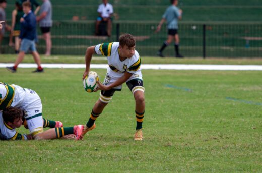 A rugby player in green and yellow prepares to pass the ball during a match on a grassy field. Teammates and spectators are visible in the background.