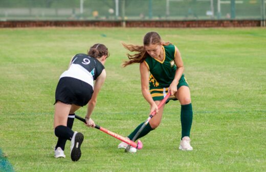 Two young women playing field hockey on a grassy field. One in green and gold defends, while the other in black and white attacks with focus.