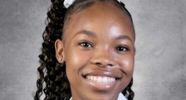 A young person smiles warmly against a neutral gray background. They have curly hair tied with a white ribbon, creating a friendly and cheerful tone.