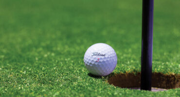 A golf ball rests on bright green grass, next to the edge of a golf hole with a flagstick. The scene conveys anticipation and precision.