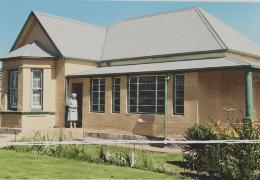 A woman stands outside a house, smiling, with a garden visible in the background.
