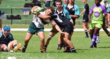 Young rugby players in action on a grassy field. A player in a white and green jersey clutches the ball tightly while being tackled by a player in black. Teammates in varied uniforms and a referee in green and purple observe intensely in the background. The scene is dynamic and intense.