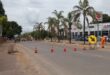 Road under construction with traffic cones and a directional sign. Workers in orange uniforms, palm trees, and car dealerships line the urban street.