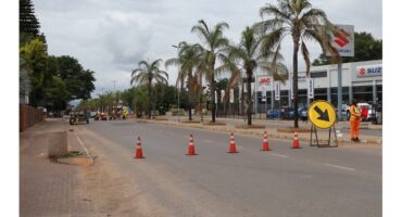 Road under construction with traffic cones and a directional sign. Workers in orange uniforms, palm trees, and car dealerships line the urban street.