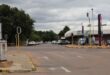 A quiet city intersection on a cloudy day, with traffic lights and parked cars lining the street. Trees and a white building flank the road.