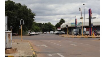 A quiet city intersection on a cloudy day, with traffic lights and parked cars lining the street. Trees and a white building flank the road.