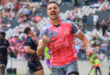 A rugby player in a pink and gray jersey celebrates energetically on the field, surrounded by teammates and spectators, conveying excitement and victory.