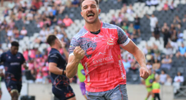 A rugby player in a pink and gray jersey celebrates energetically on the field, surrounded by teammates and spectators, conveying excitement and victory.