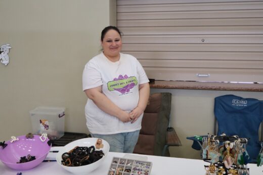 Smiling person at a craft booth with a "Jamm's Arts & Crafts" shirt. The table displays colorful handmade jewelry and accessories, creating a welcoming atmosphere.
