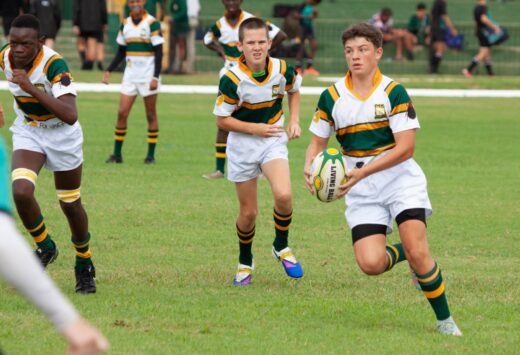 Boys in green and white rugby uniforms playing on a grassy field. One holds the ball, running determinedly as teammates follow, evoking teamwork and competition.