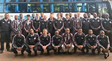 A group of young rugby players in matching black tracksuits and striped blazers pose in front of a bus with blue curtains, exuding a sense of teamwork and anticipation.