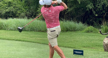 A man in a pink shirt and white hat swings a golf club on a sunny day at a golf course.