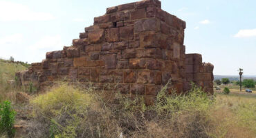 Ancient stone structure in a grassy field under a clear blue sky. The weathered blocks form a partial wall, evoking mystery and history.